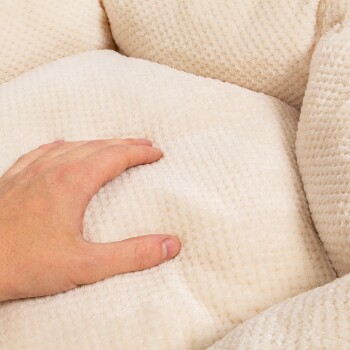 Eine Hand drückt sanft auf ein weiches, plüschiges, cremefarbenes Haustierbett mit einer strukturierten Oberfläche, die seinen Komfort und seine Qualität zeigt.