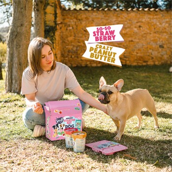 Eine Frau im Freien mit einem Französischen Bulldoggen, die eine rosa Tasche mit der Aufschrift "So-So Strawberry" und "Crazy Peanut Butter" in der Nähe von Tierfutterbehältern hält.