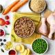 Bowl of ground meat pet food surrounded by raw chicken, carrots, peas, rice, potatoes, and various fruits on a white surface.