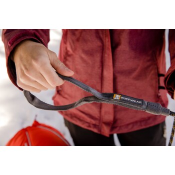 Eine Person in einer roten Jacke hält eine graue Hundeleine mit einem "Ruffwear"-Etikett, steht im Schnee mit einer roten Tasche im Hintergrund.