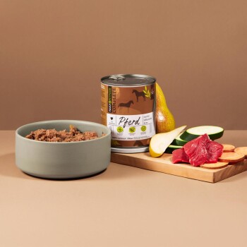 Eine Dose Pferdefleisch-Hundefutter, beschriftet "Pferd", neben einer Schüssel mit Nassfutter, mit Scheiben von Birne, Zucchini und rohem Fleisch auf einem Holzbrett.