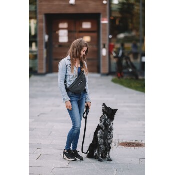 Eine Person in einer Jeansjacke und Jeans steht auf einem Steinpflaster und hält eine Leine für einen schwarz-grauen Hund, der zu ihnen aufblickt.