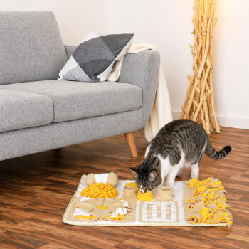 Getigerte Katze schnuppert an einer gelb-beigen interaktiven Schnüffel-Puzzlematte auf Holzboden neben einem grauen Sofa.