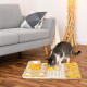 Getigerte Katze schnuppert an einer gelb-beigen interaktiven Schnüffel-Puzzlematte auf Holzboden neben einem grauen Sofa.