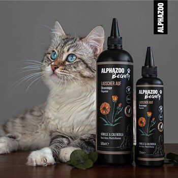 Eine flauschige Katze mit blauen Augen sitzt neben zwei Flaschen ALPHAZOO Beauty Ohrreiniger mit der Aufschrift "Läuscher Auf" und Kamille- sowie Ringelblumenmotiven.
