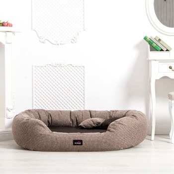 Ein gemütliches, braunes Haustierbett mit einem weichen, gepolsterten Design und einem kleinen Kissen, das in einem hellen Raum mit weißen Wänden und eleganter Dekoration platziert ist.