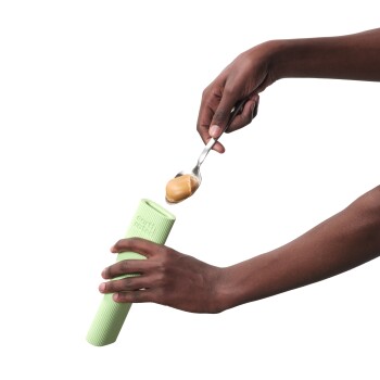 Une main tenant une cuillère de beurre de cacahuète au-dessus d'un distributeur de friandises en silicone vert, prête à le remplir pour un animal de compagnie.