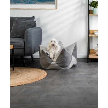 Ein kleiner weißer Hund sitzt bequem in einem stilvollen grauen Haustierbett, umgeben von moderner Möbeln und Dekor in einem gemütlichen Wohnzimmer.