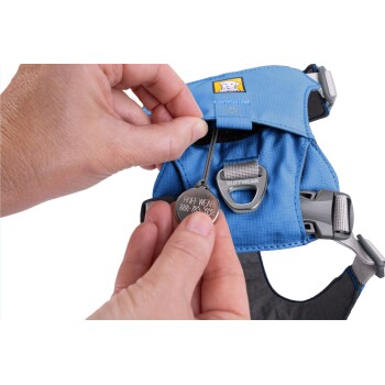 Die Hand einer Person hält ein Metalletikett mit der Aufschrift "RUFF WEAR 888-782-2922", während sie ein blaues Hundegeschirr mit einem Logo-Patch anpasst.