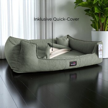 Graues Stoffhaustierbett mit Kissen und inklusive Schnellbezug, auf dunklem Boden nahe weißen Vorhängen und grüner Pflanze platziert.