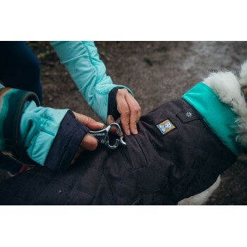 Eine Person befestigt einen Metallclip an einer schwarzen Hundejacke mit einem türkisfarbenen Kragen, die ein Ruffwear-Logo trägt.