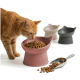 Orange gestreifte Katze schnuppert an einer pinken, erhöhten, katzenförmigen Schüssel mit Trockenfutter; Metallschaufel und zwei weitere Schüsseln im Hintergrund.
