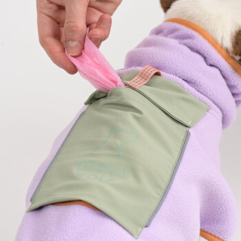 Hand zieht pinke Hundekotbeutel aus grünem Beutel mit Aufschrift „Woolly Wolf“, befestigt an einem Hund mit lila Fleecejacke.