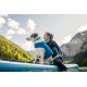 Eine Person in einem Neoprenanzug sitzt auf einem Paddleboard mit einem Hund, der eine blaue Schwimmweste trägt, umgeben von einem malerischen See und Bergen.