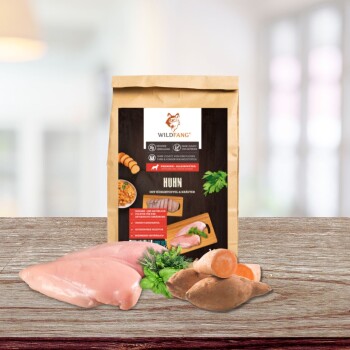 Eine Packung WILDFANG Haustierfutter mit dem Etikett 'HUHN mit Süßkartoffel & Kräutern' mit rohen Hähnchenfilets und Süßkartoffeln auf einer Holzoberfläche.