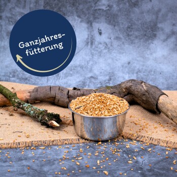 Eine Edelstahlschüssel mit Samen auf einer strukturierten Oberfläche, umgeben von Holzästen, mit "Ganzjahresfütterung" darüber.