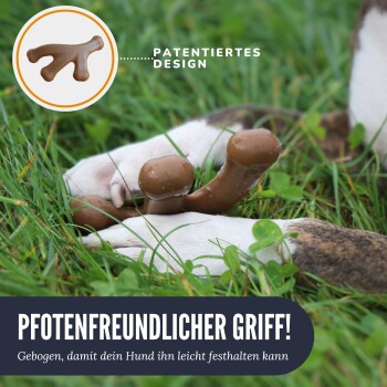 Die Pfote eines Hundes hält ein gebogenes, knochenförmiges Spielzeug auf Gras, mit dem Text 'PFOTENFRIENDLICHER GRIFF! Gebogen, damit dein Hund ihn leicht festhalten kann.'