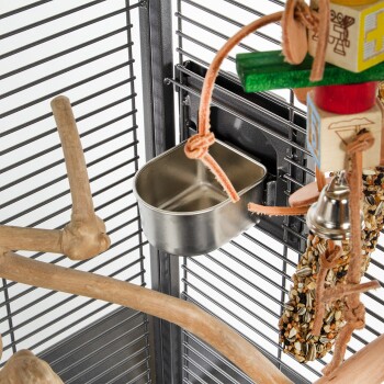 Eine Nahaufnahme eines Haustierkäfigs mit einer Futterschüssel aus Edelstahl, Holzstangen, bunten Spielzeugen und einem Saat-Leckerli, das an einem Seil hängt.