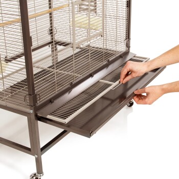 Eine Person zieht eine Schublade aus einem großen, metallenen Vogelkäfig mit Drahtgitterdesign und Holzstangen im Inneren heraus.