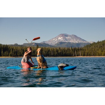 Eine Frau in einem grauen Tanktop und pinken Leggings hält ein Spielzeug auf einem blauen Paddleboard mit ihrem Golden Retriever in einem See.