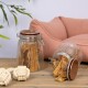 Zwei Glasbehälter mit Haustierleckereien, einer aufrecht und einer umgekippt, neben einem Seilspielzeug auf einem Holztisch, mit einem rosa Haustierbett im Hintergrund.