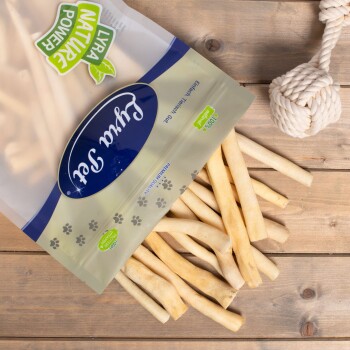 Ein Sack "Premium Qualität" Tierleckerlis mit einem "100% Natürlich"-Siegel, der leichte Kausticks neben einem weißen Seilspielzeug auf Holz auskippt.