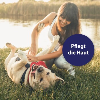 Eine Frau in einem weißen Tanktop spielt mit einem glücklichen gelben Labrador auf grünem Gras am Wasser, mit einer Sprechblase, die sagt "Pflegt die Haut."