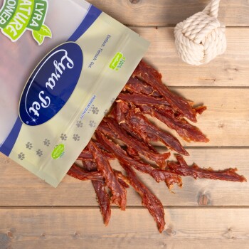 Eine Tüte Haustierleckerlis mit der Aufschrift "Fruity Snack", die Streifen von getrocknetem Fleisch auf einer Holzoberfläche auskippt, neben einem geknoteten Seilspielzeug.