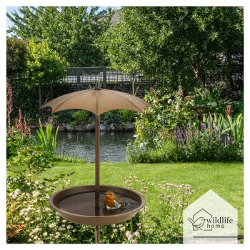 Ein Vogelbad mit einem kleinen Vogel, der sich unter einem beigen Schirm badet, umgeben von einem üppigen Garten und einem Teich im Hintergrund.