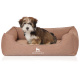 Ein kleiner, flauschiger Hund sitzt bequem in einem gemütlichen braunen Hundebett mit dem Logo "KNUFFELWUFF", das auf der Vorderseite angezeigt wird.