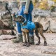 Eine Person in einem grauen Pullover und blauen Jeans führt einen gestromten Hund, der ein blaues Geschirr und schützende Booties auf felsigem Terrain trägt.
