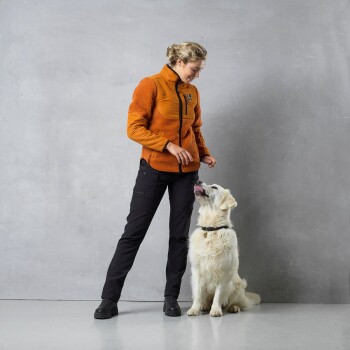 Eine Frau in einer orangefarbenen Fleecejacke steht neben einem Golden Retriever, der zu ihr aufblickt. Der Hintergrund ist eine schlichte graue Wand.