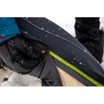 Nahaufnahme einer Person in einem schwarzen Handschuh, die eine Hundejacke mit einem grünen Reißverschluss schließt, umgeben von fallendem Schnee.