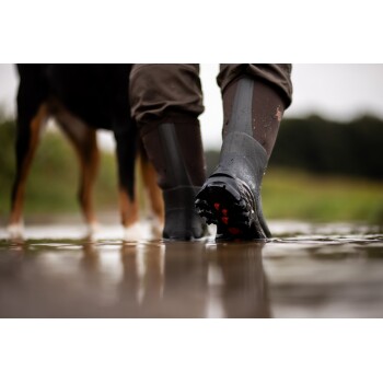 Nahaufnahme einer Person mit wasserdichten Stiefeln, die durch eine Pfütze geht, mit einem Hund im Hintergrund im Freien.