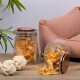 Ein Glasbehälter, gefüllt mit Haustierleckereien, wird gezeigt, mit einigen Leckereien, die herausgefallen sind. Ein plüschiges pinkes Haustierbett und ein weißes Seilspielzeug sind in der Nähe.