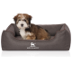 Ein kleiner, flauschiger Hund sitzt bequem in einem grauen Haustierbett mit braunem Rand, mit dem Logo "KNUFFELWUFF" deutlich sichtbar.