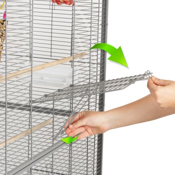 Eine Person entfernt ein Metallgitter aus einem Haustierkäfig mit grünen Pfeilen, die die Richtung zeigen. Der Käfig hat Holzstangen und einen Futternapf.