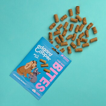 Eine Tüte Edgard Cooper Top Dog Bites auf einem türkisfarbenen Hintergrund, mit hundeförmigen Leckerlis und verspielten Hund- und Katzenillustrationen.