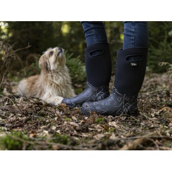 Eine Person in stilvollen schwarzen und gemusterten Gummistiefeln steht im Wald, während ein flauschiger Hund zu ihnen aufblickt.