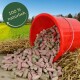 Roter Eimer, der pinke und grüne Kleintierpellets auf Heu in einem Feld verschüttet; grünes Siegel mit der Aufschrift '100% natürlich'.