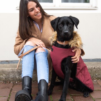 Eine Frau in einem beigen Mantel sitzt auf einer Steinmauer mit einem schwarzen Hund, der einen roten Mantel mit einer gefütterten Kapuze trägt.