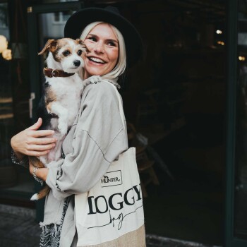 Eine Person, die einen beigen übergroßen Pullover und einen schwarzen Hut trägt, hält einen kleinen Hund nah bei sich, während sie eine Tragetasche mit der Aufschrift "HUNTER DOGGY" trägt.