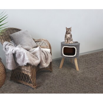 Ein stilvolles graues Katzenbett mit einem gemütlichen Innenraum steht auf Holzbeinen, während eine getigerte Katze oben sitzt, umgeben von einem Korbstuhl und einer weichen Decke.