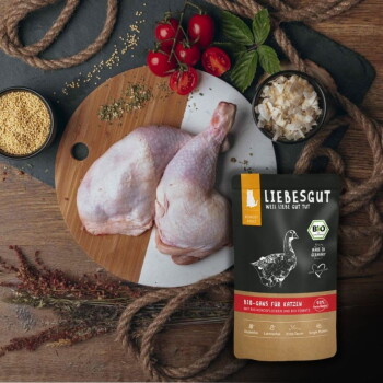 LIEBESGUT Katzenfutterbeutel (Bio-Gans für Katzen) neben rohen Geflügelschenkeln, Cherrytomaten und Körnern auf einem hölzernen Schneidebrett.