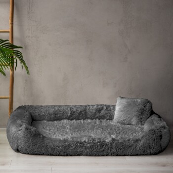 Ein plüschiges, hellgraues Haustierbett mit erhöhten Rändern und einem kleinen Kissen, vor einer strukturierten Wand und Holzdekoration.