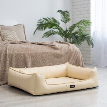 Ein gemütliches, beiges Haustierbett mit erhöhten Rändern steht auf einem hellen Holzboden, neben einem Bett, das mit einer braunen Decke bedeckt ist, und einer grünen Pflanze im Hintergrund.