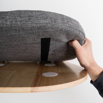 Eine entspannte grau-weiße Katze mit auffälligen Augen liegt auf einem runden, grau bezogenen Platz, der an einer Wand befestigt ist.