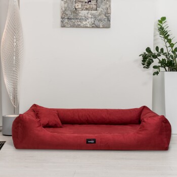 Rote Haustierbett mit weichem Stoff und erhöhten Kanten, ausgestattet mit einem kleinen schwarzen Etikett, in einem modernen Raum mit einer Pflanze und einer dekorativen Lampe.