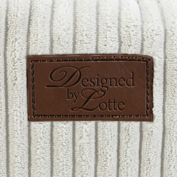 Gros plan d'un lit pour animaux présentant un tissu crème texturé avec des rayures grises et une étiquette en cuir marron portant la mention "Conçu par Lotte."