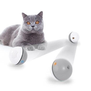 Eine graue Katze mit orangefarbenen Augen liegt neben einem weißen Haustierspielzeug, das einen Lautsprecher und eine Lichtanzeige hat.